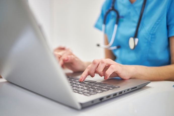 Close-up hands of unrecognizable clinician in medical uniform working typing on laptop keyboard sitting at desk